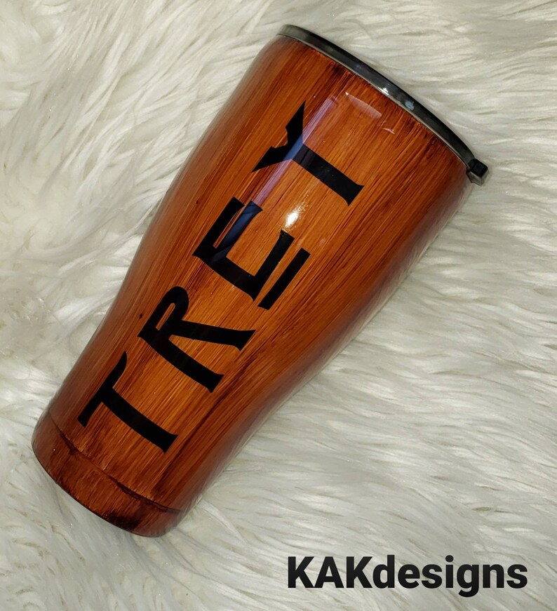 May include: A brown and black stainless steel tumbler with the name "TREY" printed on the side. The tumbler has a wood grain design.