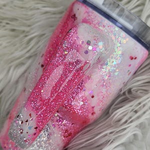 Puede incluir: Un vaso cubierto de purpurina con asa, con un dise&ntilde;o de m&aacute;rmol rosa y blanco. El asa y el cuerpo est&aacute;n adornados con purpurina rosa y plateada, con una tapa transparente. El vaso est&aacute; dise&ntilde;ado para bebidas fr&iacute;as.