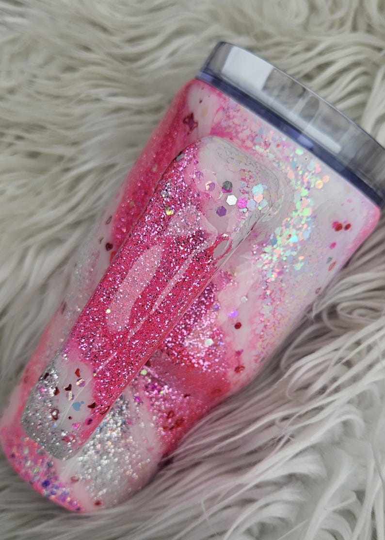 Puede incluir: Un vaso cubierto de purpurina con asa, con un dise&ntilde;o de m&aacute;rmol rosa y blanco. El asa y el cuerpo est&aacute;n adornados con purpurina rosa y plateada, con una tapa transparente. El vaso est&aacute; dise&ntilde;ado para bebidas fr&iacute;as.