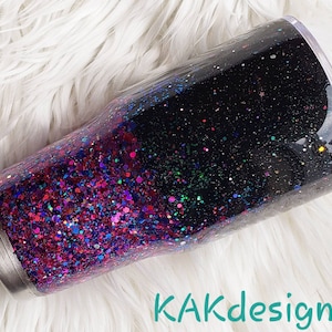 May include: A black and pink glitter tumbler with a clear lid. The tumbler is filled with a mix of pink, purple, and blue glitter. The glitter is layered in a gradient from pink at the bottom to black at the top. The tumbler is sitting on a white fluffy surface. KAKdesigns is written in teal text at the bottom of the image.