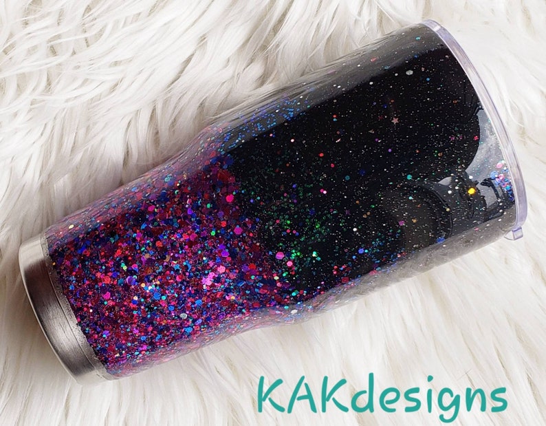 May include: A black and pink glitter tumbler with a clear lid. The tumbler is filled with a mix of pink, purple, and blue glitter. The glitter is layered in a gradient from pink at the bottom to black at the top. The tumbler is sitting on a white fluffy surface. KAKdesigns is written in teal text at the bottom of the image.