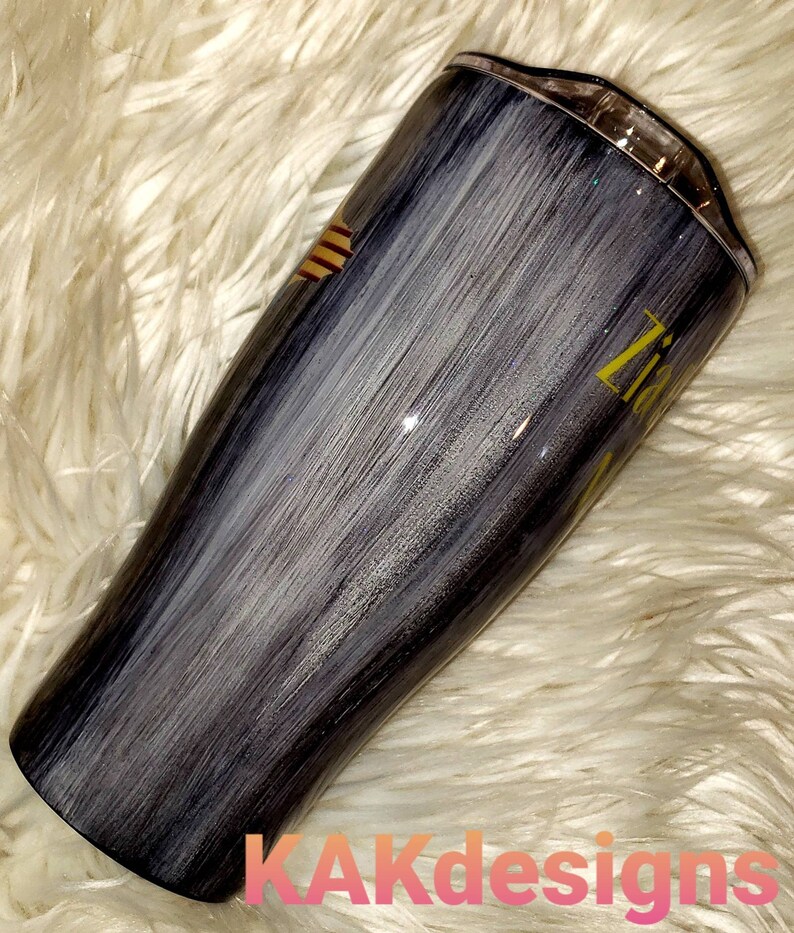 May include: A black and white stainless steel tumbler with a gold glitter finish. The tumbler has a brushed metal look and a gold stripe design. The text "Zita" is written in gold on the side of the tumbler.