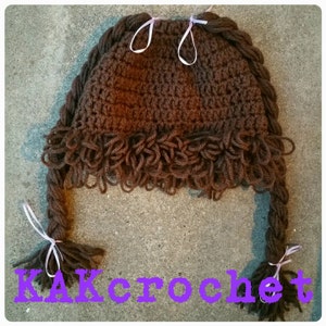Puede incluir: Un gorro marrón tejido a ganchillo con dos largas trenzas, cada una atada con una cinta blanca. El gorro tiene un diseño texturizado y una franja de hilo alrededor de la parte inferior. El texto "KAKcrochet" es visible en morado.