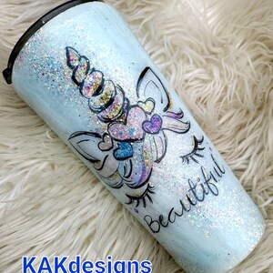 May include: A light blue tumbler with a unicorn design and glitter. The unicorn has a rainbow horn and pink hearts. The tumbler says "Beautiful" on the side.