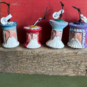 May include: Four hand-painted wooden ornaments shaped like figures with white beards, each featuring a black hanging string and a small white price tag. The figures are painted in blue, red, light blue, and purple, with decorative buttons on top.