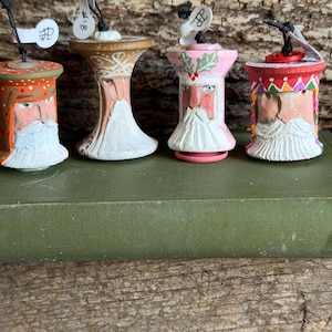 May include: Four hand-painted wooden spool ornaments featuring bearded figures. Each spool has a black hanging string and a small white price tag. The figures include varied hat colors like orange, brown, pink, and red, with detailed facial features and white beards.