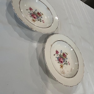 Puede incluir: Dos cuencos de cerámica blanca con un borde dorado y un patrón floral. Los cuencos tienen un borde festoneado y están decorados con flores rosas, amarillas y azules.