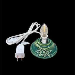 May include: A green ceramic Christmas tree light base with a clear bulb. The base has a holly leaf design and a white cord with a plug. The light is designed for use with a Christmas tree.