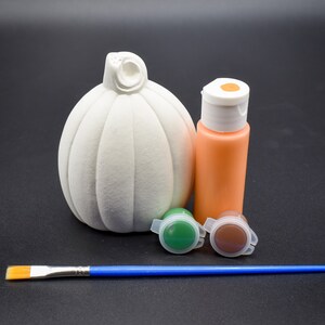 May include: A white ceramic pumpkin with a stem, a small bottle of orange paint, two small bottles of green and brown paint, and a blue paintbrush.