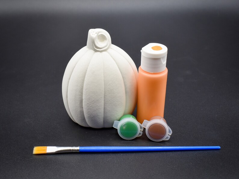 May include: A white ceramic pumpkin with a stem, a small bottle of orange paint, two small bottles of green and brown paint, and a blue paintbrush.