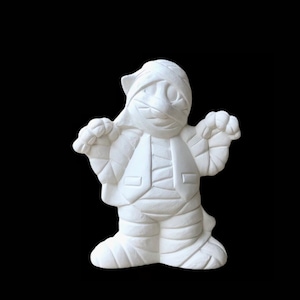 May include: White ceramic mummy figurine with a bandage-wrapped body and a smiling face. The mummy is standing with its arms outstretched, as if in a welcoming gesture.