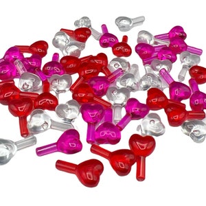 Puede incluir: Una colección de cubiertas de bombillas transparentes en forma de corazón en rojo, rosa y transparente. Cada cubierta de bombilla tiene un diseño en forma de corazón y un pequeño tallo. Las bombillas están dispersas sobre un fondo blanco.