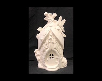 May include: White ceramic fairy house with a door, a window, and a dragonfly on the roof. The house is decorated with leaves and other natural elements.