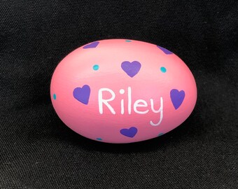 May include: Pink painted egg with purple hearts and turquoise dots. The name "Riley" is written in white paint.