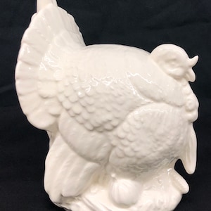 May include: White ceramic turkey figurine with detailed feathers and a textured surface. The turkey is in a seated position with its head turned to the side.