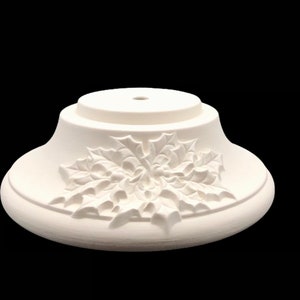 May include: White ceramic Christmas tree stand with a holly design. The stand has a round top with a hole in the center and a wider base with a raised holly design.