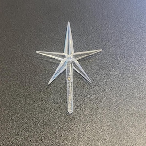 May include: A clear, five-pointed star-shaped ornament with a textured stem. The star is made of transparent plastic and is designed to be placed on top of a Christmas tree. The ornament is shown against a dark background.
