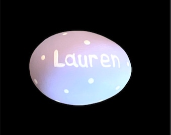 May include: A light purple painted rock with white polka dots and the name "Lauren" written in white.