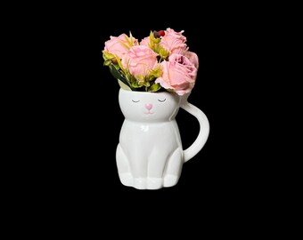 May include: White ceramic cat planter with a pink nose and closed eyes, holding a bouquet of pink roses.