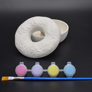 May include: A white ceramic donut-shaped container with a matching small bowl, and a set of paint pots in pink, purple, yellow, and blue. A blue paintbrush is also included, all on a black background.