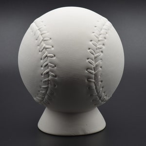 May include: White ceramic baseball bank with raised stitching detail. The bank is on a small base.