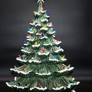 May include: A green ceramic Christmas tree with white faux snow and colorful lights. The tree is lit and has a star on top.