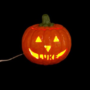 May include: A lit-up orange ceramic pumpkin with a carved jack-o'-lantern face and the word "LUKE" carved inside.