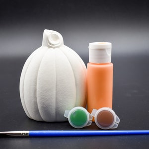May include: A white ceramic pumpkin with a stem, a small bottle of orange paint, two small pots of green and brown paint, and a blue paintbrush.