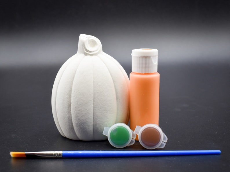 May include: A white ceramic pumpkin with a stem, a small bottle of orange paint, two small pots of green and brown paint, and a blue paintbrush.