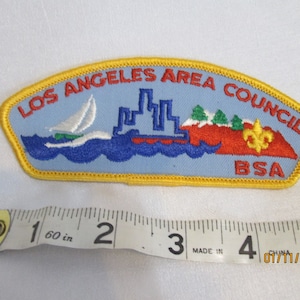 May include: Embroidered patch with the text "LOS ANGELES AREA COUNCIL BSA". The patch features a sailboat, city skyline, mountains, and a fleur-de-lis. The patch is blue, red, green, yellow, and white.