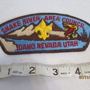 May include: Embroidered patch with the text "SNAKE RIVER AREA COUNCIL IDAHO NEVADA UTAH". The patch features a mountain range, a yellow fleur-de-lis, and a snake. The patch is approximately 10 cm long.