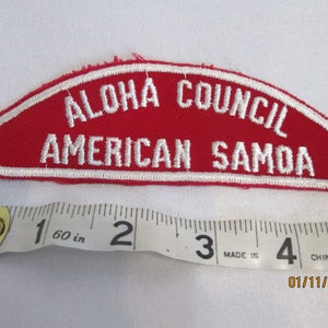 Op de afbeelding: Een rood en wit geborduurde patch met de tekst "ALOHA COUNCIL AMERICAN SAMOA". De patch heeft een gebogen rechthoekige vorm. Een meetlint is eronder geplaatst.
