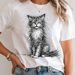 Könnte beinhalten: Weißes T-Shirt mit einer detaillierten Schwarz-Weiß-Illustration einer flauschigen Katze mit langem Fell und Wassertropfen. Die Katze sitzt mit einem leicht mürrischen Gesichtsausdruck. Ein lässiges Kleidungsstück für den Alltag.