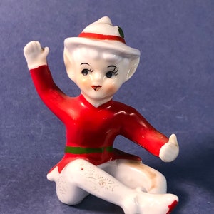 May include: A small, white porcelain figurine of an elf wearing a red and white hat, a red shirt with a green belt, and white trousers. The elf is sitting with one hand raised and the other resting on its lap.