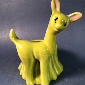 May include: A green ceramic deer planter with a pink nose and black eyes. The deer is standing on a green base with a ruffled skirt.