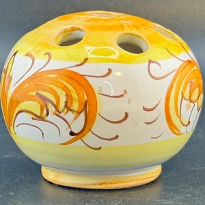 May include: A ceramic, spherical vase with a yellow and white color scheme. The vase features orange floral designs and three holes on top. The base is a light yellow color.