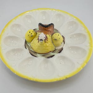 May include: A vintage ceramic egg serving platter with a yellow rim and a central decorative feature. The centre has a yellow hen and two yellow chicks in a nest. The platter has 12 egg-shaped indentations.