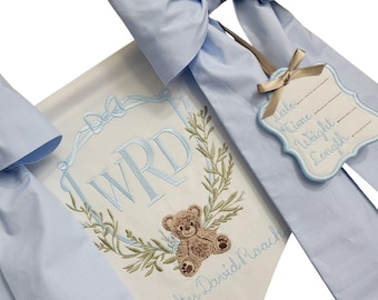 May include: A light blue banner with a large bow, featuring embroidered details. The banner displays the initials "WRD" within a wreath, a teddy bear, and the name "Walter David Roach". A tag with space for date, time, weight, and length is attached.