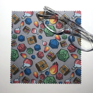 May include: A gray microfiber cleaning cloth with a repeating pattern of dice, treasure chests, potion bottles, and flames. The cloth is designed for cleaning glasses.