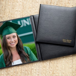 May include: A black leather diploma cover with gold lettering that reads "Jennifer Baltimore Class of 2023". The cover is open to reveal a photo of a woman in a green graduation gown and cap.
