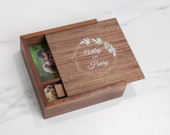 May include: A wooden box with a hinged lid. The lid is open and shows a photo of a couple inside the box. The lid is engraved with a gold geometric frame and a floral design with the text "Ashley and Henry".