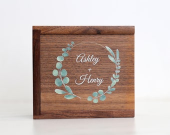 May include: A wooden box with a light brown finish. The lid features a wreath of green leaves with the names "Ashley + Henry" written in white inside the wreath.