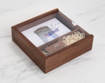 May include: A wooden box with a clear lid, containing a photograph, a USB drive, and decorative straw. The box has a dark brown finish. The USB drive is engraved with initials. The photograph has the text "Mae Cohen photography".