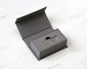 May include: A gray gift box with a hinged lid, open to reveal a smaller gray box containing a silver USB drive. The USB drive has the initials "A-H" etched on it. The background is a white marble surface.