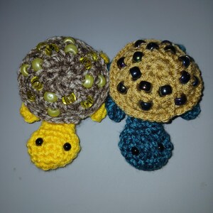 May include: Two crocheted turtle figures. The left turtle has a brown shell with yellow beads and a bright yellow head. The right turtle has a tan shell with dark metallic beads and a teal head. Both have small black bead eyes and four flippers.