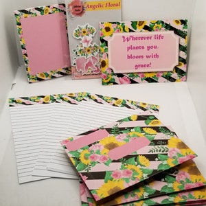 May include: A set of stationery products featuring a floral design with sunflowers and roses on a pink and black striped background. The set includes note cards, envelopes, and a blank card with a pink frame. The text on the card reads "Wherever life plants you, bloom with grace!"