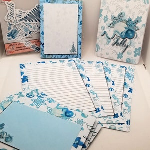 May include: A set of blue and white Christmas stationery featuring a snowflake and ornament design. The set includes a notepad, notecards, and envelopes. The notecards have lined paper and the envelopes are blank. The notepad has a blue background with a white snowflake design. The notecards and envelopes have a white background with a blue snowflake and ornament design. The text "Peace Love Joy" is printed on the notecards and envelopes.