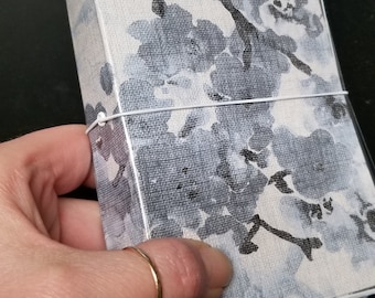 May include: A floral patterned journal with a clear plastic cover. The cover features a design of gray and black flowers and branches on a light gray background. A thin, white elastic band wraps around the journal.