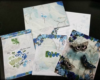 May include: A set of blue and white stationery featuring floral and butterfly designs. The set includes envelopes, note cards, and lined paper. The text "The lan Studio Stationery Shoppe" is visible on one of the envelopes.