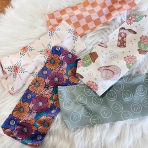 May include: Five different patterned headbands. One headband has a pink and orange checked pattern. Another headband has a floral pattern with pink, orange, and purple flowers. Another headband has a white background with a floral pattern and a cactus. Another headband has a white background with a floral pattern and a boot. The last headband has a green background with a white smiley face pattern.